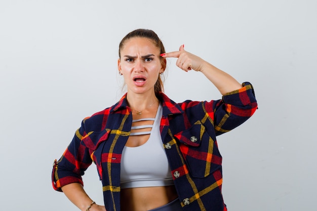 Young female in crop top, checkered shirt pointing at head and looking puzzled , front view.