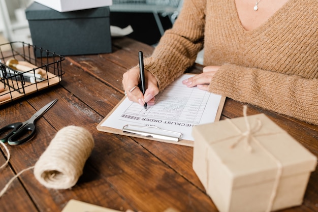 Young female customer in beige knitted pullover sitting by wooden table and putting ticks opposite ordered goods in checklist