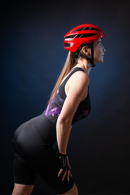 A young female cyclist wearing a safety helmet and glasses dressed in a bib shorts poses against a black background in the studio