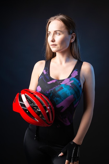 A young female cyclist wearing a safety helmet and glasses dressed in a bib shorts poses against a black background in the studio