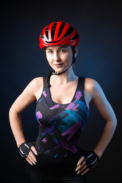 A young female cyclist wearing a safety helmet and glasses dressed in a bib shorts poses against a black background in the studio