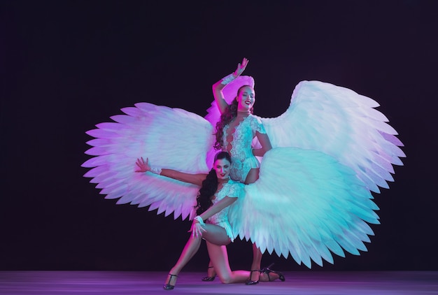 Young female dancers with white angel's wings in neon colors. Graceful models, women dancing, posing.