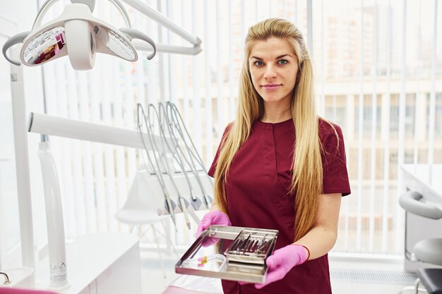 Young female dentist in uniform standing in stomatology office with tools in hands