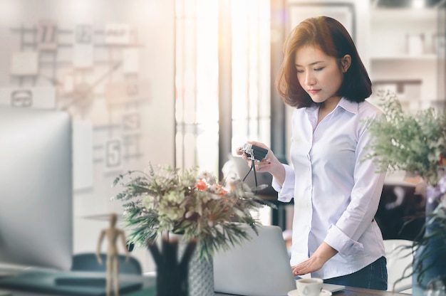 Giovane designer femminile nella macchina fotografica della tenuta di abbigliamento casual e nel funzionamento del computer portatile