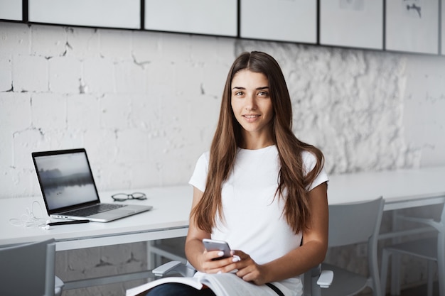 Giovane designer femminile che utilizza un computer portatile e un telefono cellulare che legge una rivista fresca per l'ispirazione.