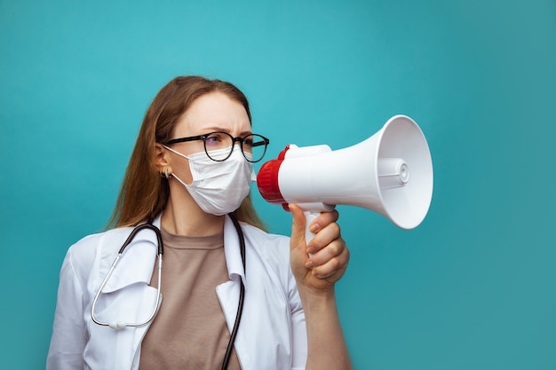 Young female doctor in face mask shouting into megaphone on blue.