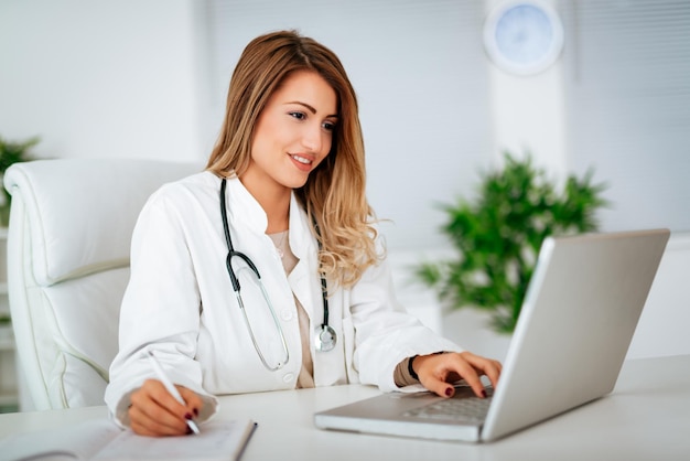 Young female doctor in her consulting room, writing on laptop and checking medical records.