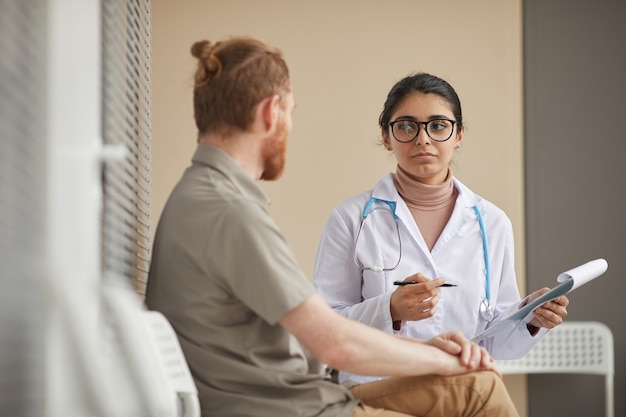 Фото Молодая женщина-врач держит медицинскую карту и обсуждает с пациентом его болезнь, сидя в коридоре