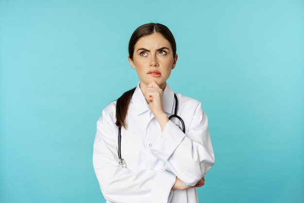 Young female doctor, hospital worker in white coat, thinking and looking away thoughtful, searching solution, standing over toquoise background.