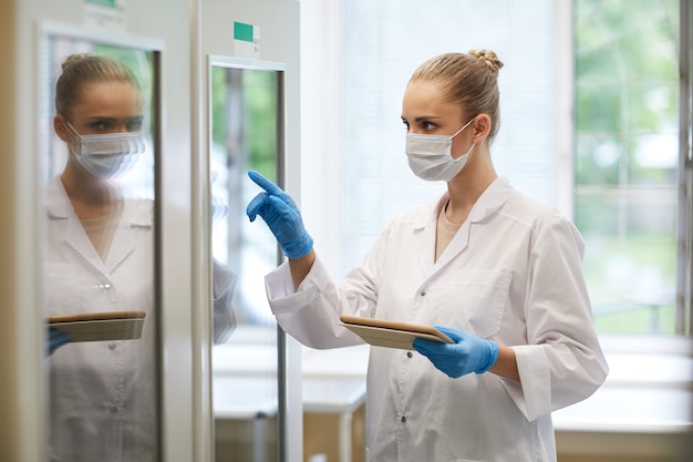 Young female doctor in mask and in white coat using digital tablet at work she pointing at fridge while working at the lab