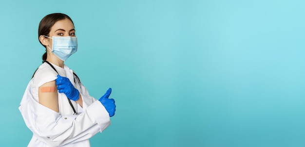 Young female doctor nurse in medical face mask and hospital uniform showing thumbs up after getting covid19 omicron vaccine showing patch on shoulder blue background