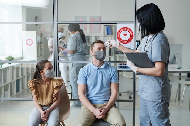 Young female doctor or nurse in uniform measuring body temperature of man in protective mask and casualwear sitting next to his daughter