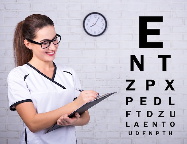 Young female doctor ophthalmologist with eye test chart over white brick wall background