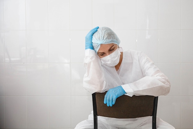 A young female doctor in a protective suit cap mask and gloves is sitting tired on a chair in the me...