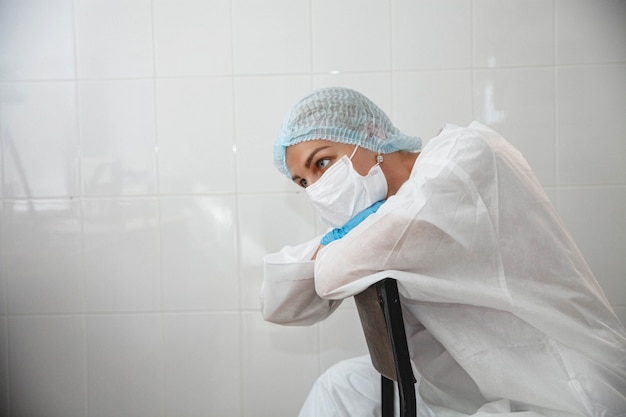 A young female doctor in a protective suit cap mask and gloves is sitting tired on a chair in the me...