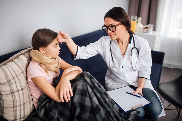 Il giovane medico femminile si siede accanto al bambino malato. tiene la mano sulla fronte del bambino. guarda la bambina e sorride. la ragazza stanca si siede sullo strato in una stanza con il medico.