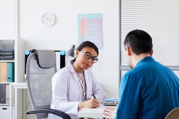 Young female doctor sitting at the table making notes and listening to patient during his visit at office