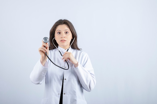 Young female doctor using stethoscope to check pulse on white wall.