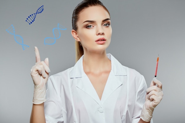 Young female doctor wearing white medical robe and holding syringe at gray background, health care and pharmacology concept, girl in white uniform pointing at DNA model