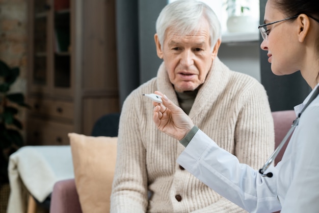 Young female doctor in whitecoat looking at thermometer after measuring body temperature of sick senior man sitting on couch in living-room