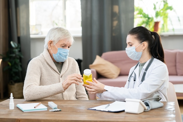 Young female doctor in whitecoat and protective mask holding bottle of pills over table, showing and prescribing them to sick senior man