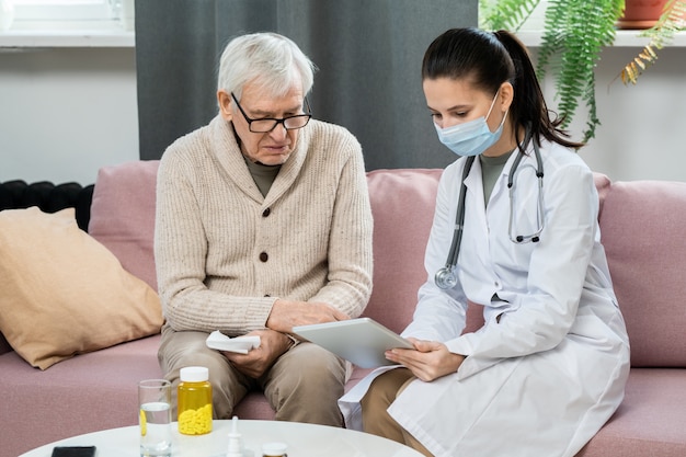 Young female doctor in whitecoat and protective mask holding digital tablet while sitting on couch next to sick senior man and consulting him