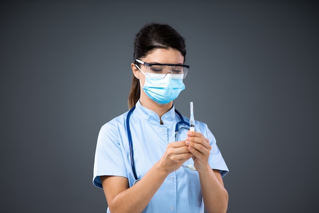 Young female doctor with face mask and protective glasses on holding syringe and preparing to give a shot to a sick patient.