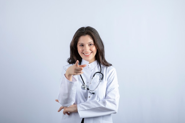 白い壁にポーズをとる聴診器を持つ若い女性医師。