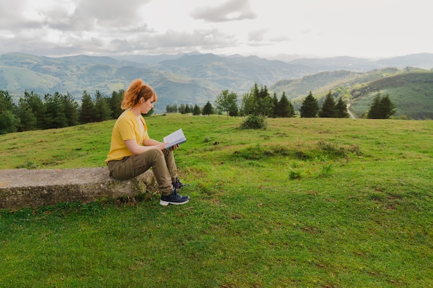 Young female dressed in yellow t-shirt sitting on a stone reading a book in the green field on the background of the mountains with a cloudy sky