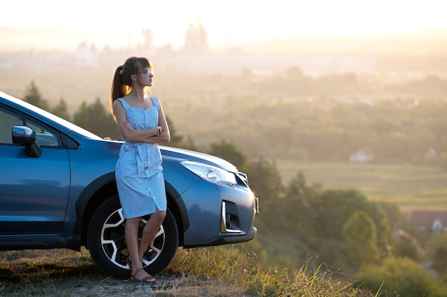 Young female driver resting near her car enjoying warm summer evening. Travel and getaway concept.