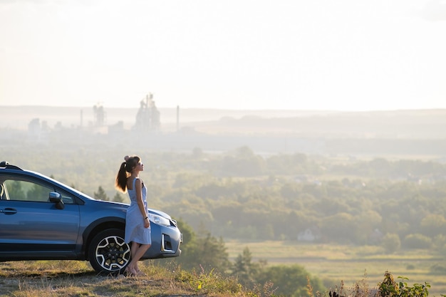 Young female driver resting near her car enjoying warm summer evening. Travel and getaway concept.