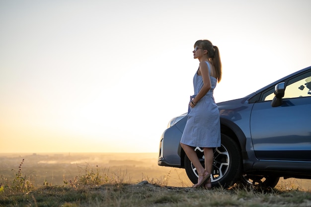 暖かい夏の夜の旅行と休暇のコンセプトを楽しんでいる彼女の車の近くで休んでいる若い女性ドライバー