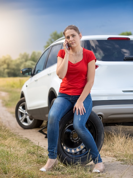 Foto giovane autista donna seduta accanto all'auto rotta al campo e parlando al telefono