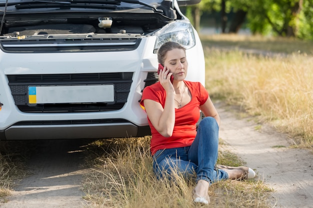 Young female driver sitting on ground next to broken car and calling for help