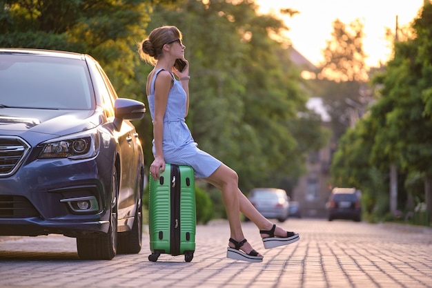 Young female driver sitting on suitcase near her car speaking on her sellphone on a city street in summer.