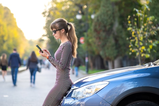 Young female driver standing near her car talking on mobile phone on a city street in summer.