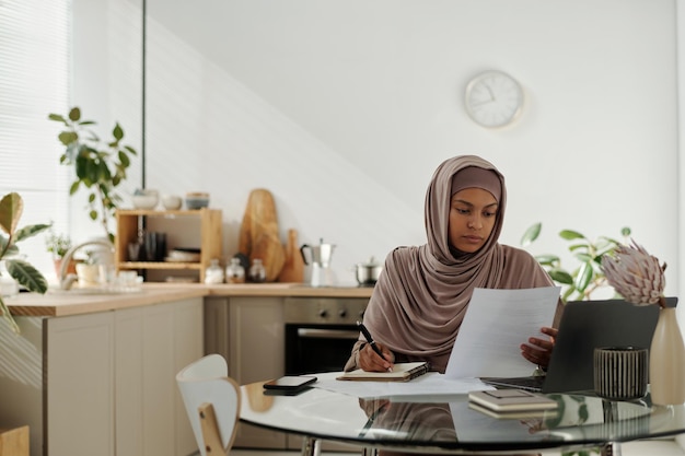 Young female employee reading financial document or contract and making notes