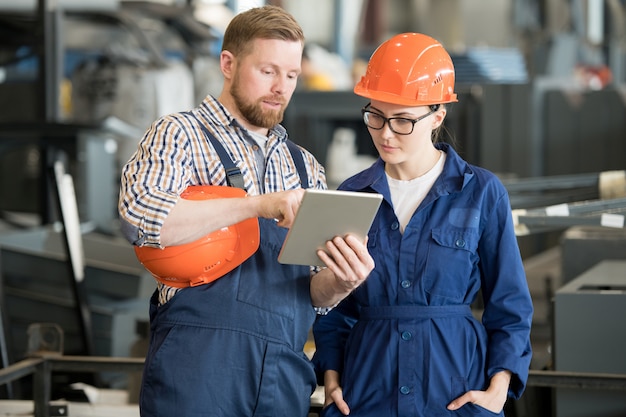 Young female engineer in uniform and helmet looking at tablet display while listening to colleague explanation of data