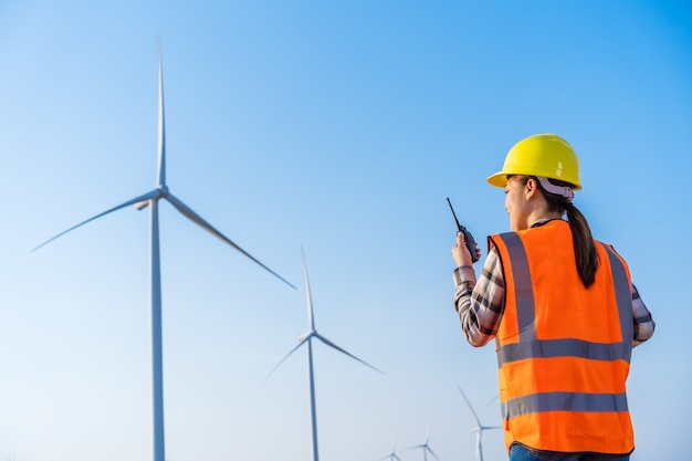 Young female engineer using walkie talkie to checking system against wind turbine farm