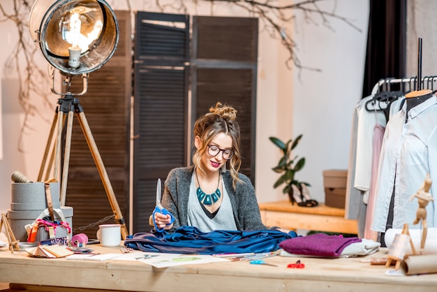 Foto giovane stilista femminile che lavora con tessuto blu seduto nel bellissimo ufficio con diversi strumenti di sartoria sul tavolo