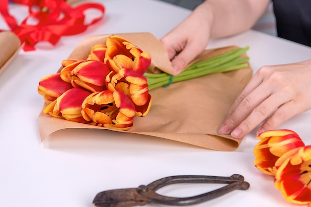 La giovane fiorista sta avvolgendo un bouquet di fiori di tulipano di colore rosso per il regalo per la festa della mamma con fiocco di nastro cravatta su uno sfondo bianco da tavolo primo piano