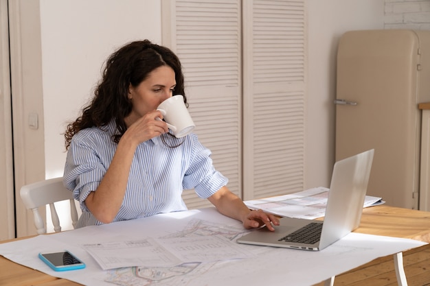 若い女性のフリーランサーがノートパソコンでリモートでテーブルワークでコーヒーを飲む