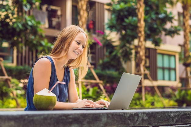 Young female freelancer sitting near the pool with her laptop in the hotel browsing in her smartphone. Busy at holidays. Distant work concept. Copy space for your text