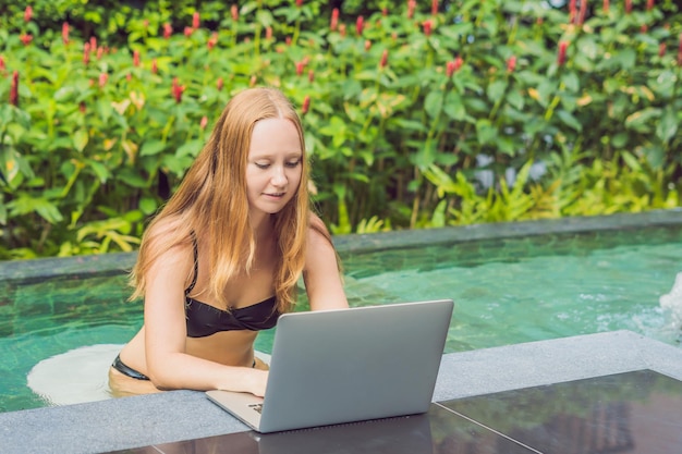 Young female freelancer sitting near the pool with her laptop in the hotel browsing in her smartphone. Busy at holidays. Distant work concept. Copy space for your text