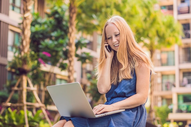 Young female freelancer sitting near the pool with her laptop in the hotel browsing in her smartphone. Busy at holidays. Distant work concept. Copy space for your text.