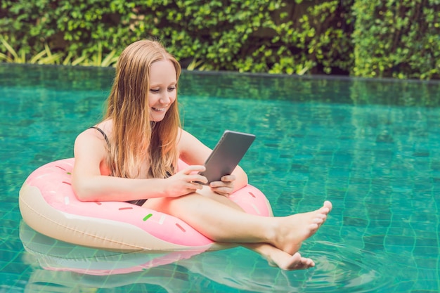 Young female freelancer sitting near the pool with her laptop in the hotel browsing in her smartphone. Busy at holidays. Distant work concept. Copy space for your text.