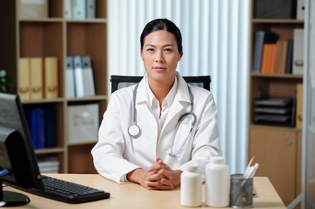 Young female general practitioner in lab coat sitting by workplace in clinics