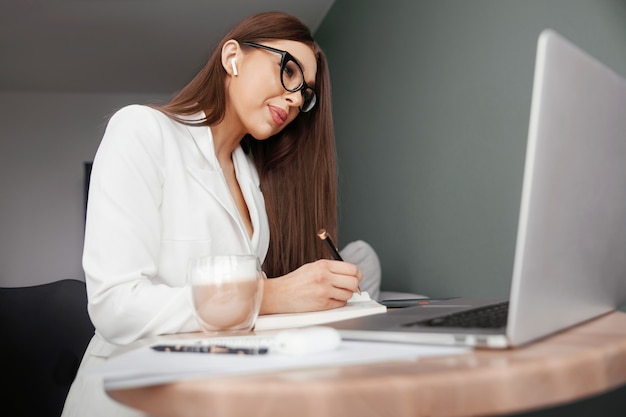 Young female in glasses using laptop, communicates on internet with customer in home, coffee mug on table.Cozy office workplace, remote work, Elearning concept