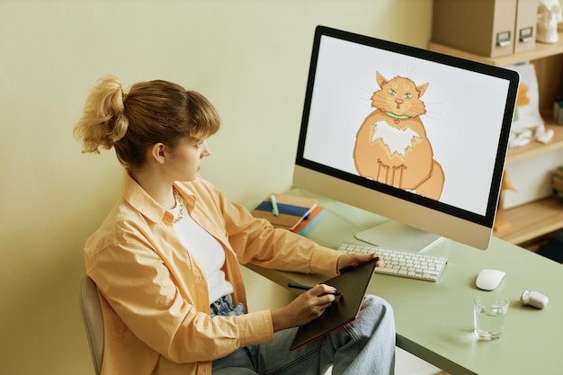 캐주얼웨어를 입은 젊은 여성 그래픽 디자이너는 태블릿 화면에 뚱뚱한 고양이를 그리고 디지털을 보고 있습니다.