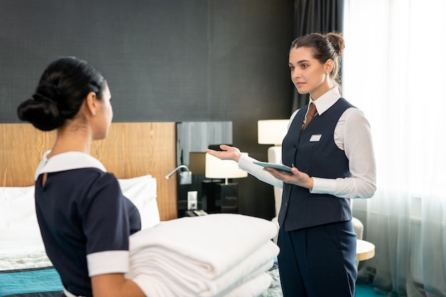 Young female head waiter in uniform talking to chambermaid in bedroom